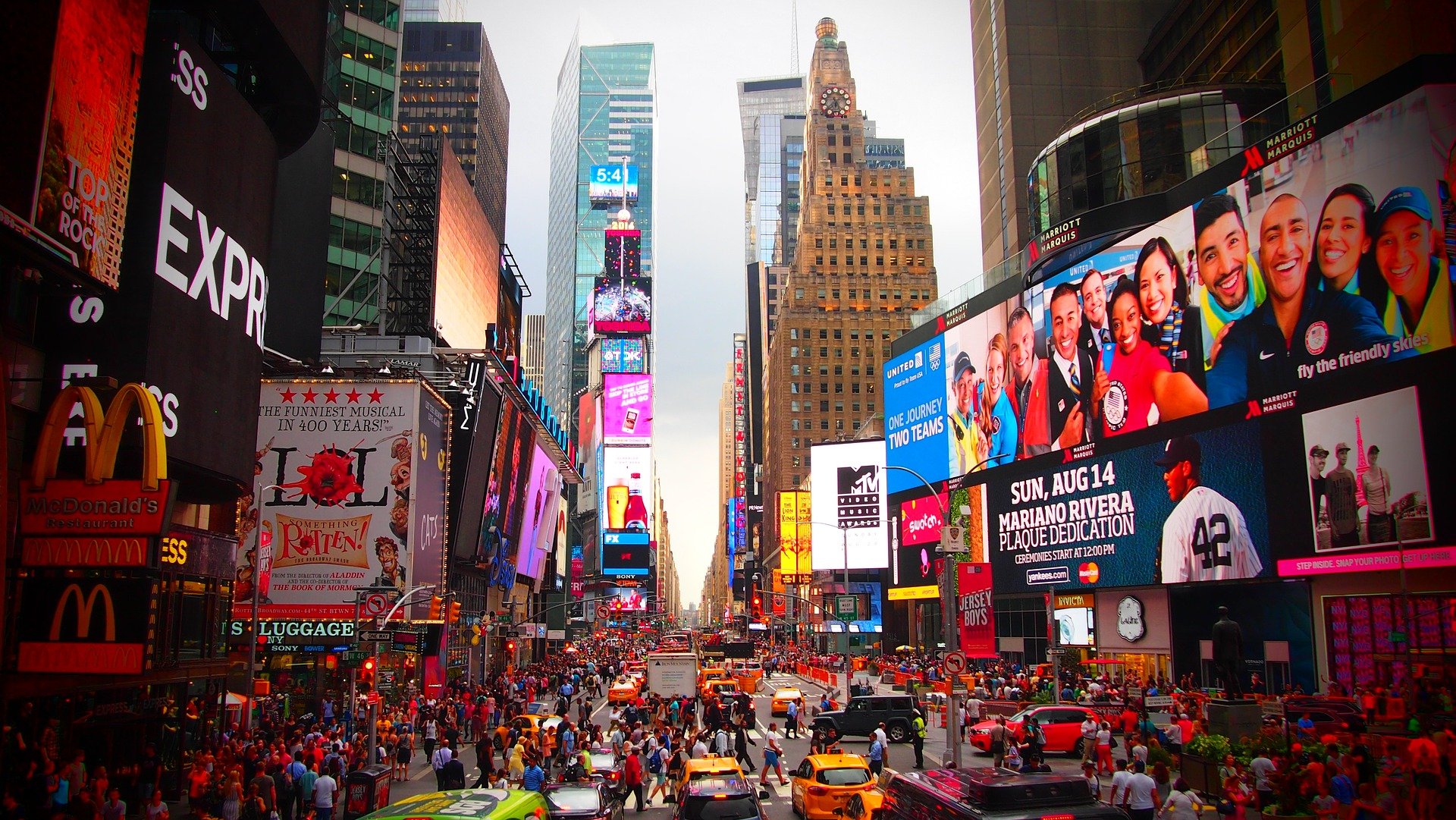 A wide view of Times Square in New York City filled with crowds of pedestrians and yellow taxis moving through the busy intersection. Bright digital billboards and towering screens line both sides of the street.